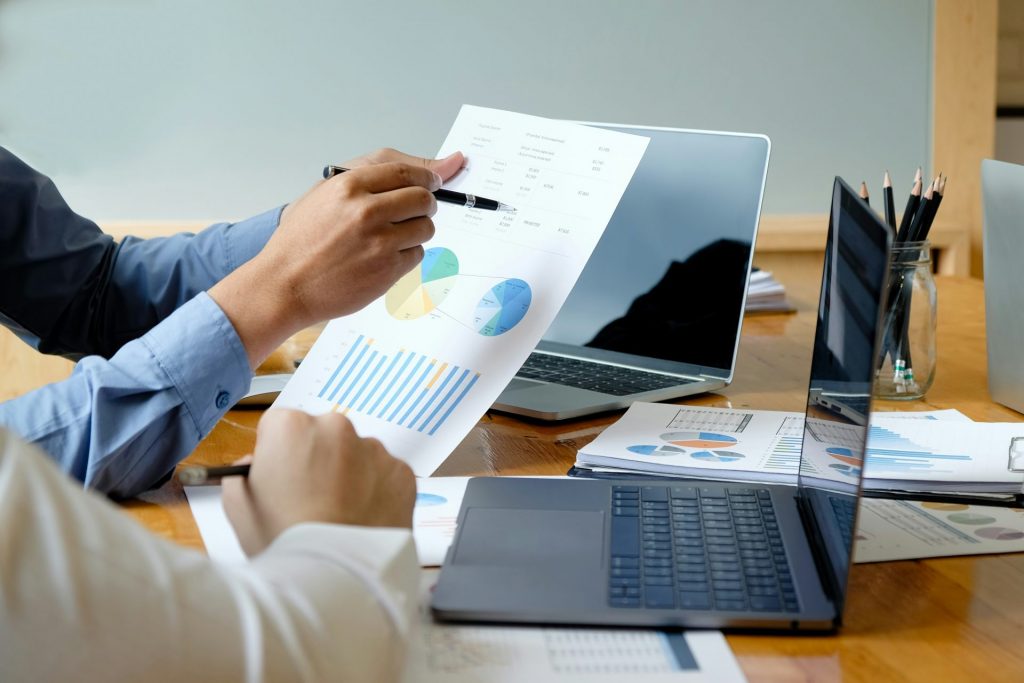 Zwei Personen analysieren Finanzunterlagen und Diagramme am Laptop – Symbolbild für die neuen Anforderungen an eine detaillierte und prüffähige Kostenrechnung im Zuge der Neuregelung des Gemeinkostenzuschlags.