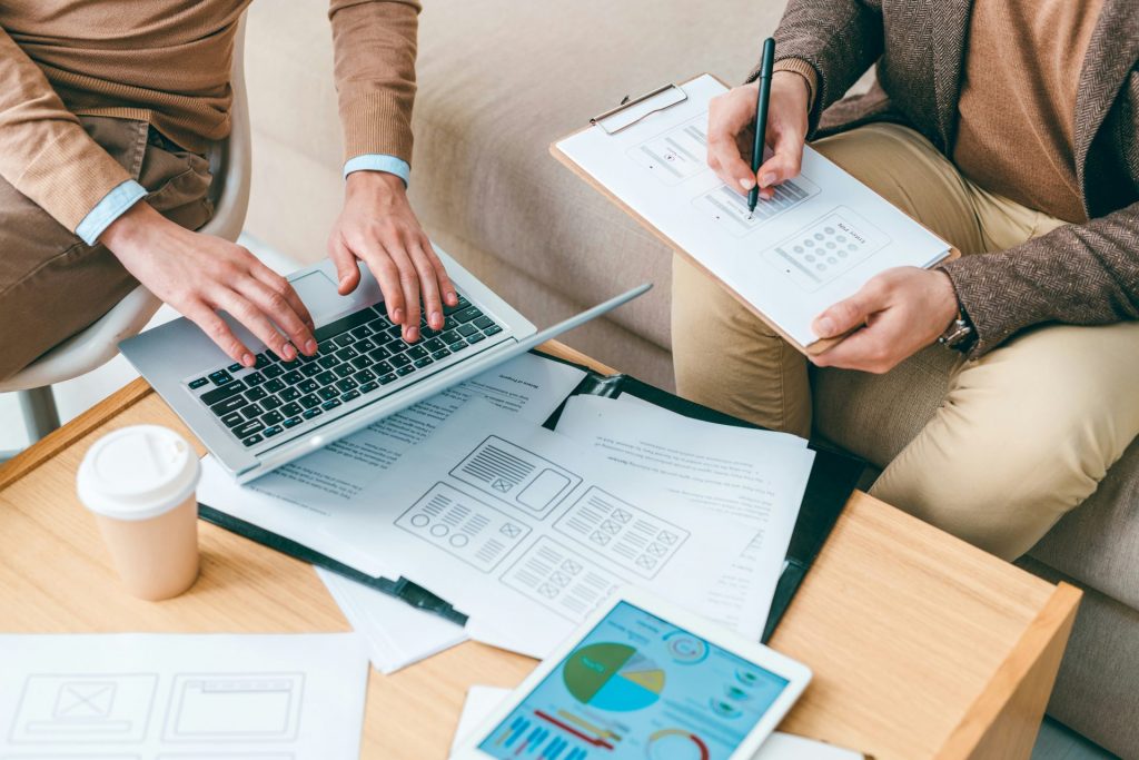 Zwei Fachkräfte arbeiten gemeinsam an einem Tisch, um Konzepte zur energieeffizienten Anlagentechnik zu analysieren. Während eine Person Daten auf einem Laptop überprüft, notiert die andere Informationen auf einem Klemmbrett. Auf dem Tisch befinden sich technische Dokumente mit Diagrammen und Tabellen sowie ein Tablet mit einem farbigen Energie-Diagramm – Hinweise auf eine detaillierte Planung oder Optimierung technischer Anlagen im Sinne der Energieeffizienz.