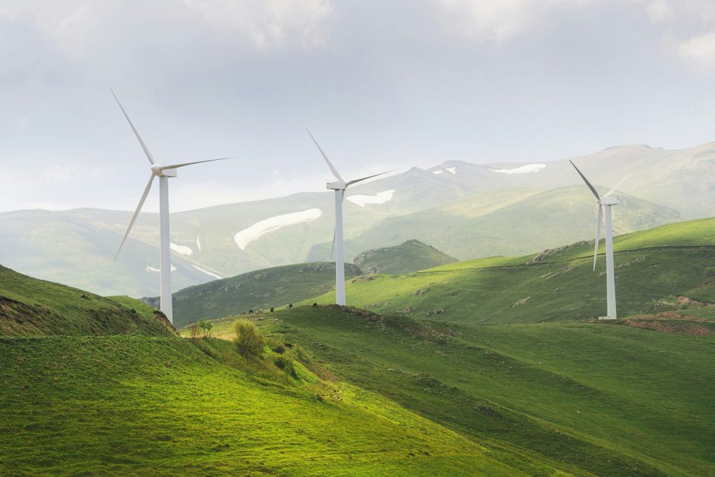 Eine grüne Hügellandschaft mit drei modernen Windkraftanlagen, die sich harmonisch in die Natur einfügen. Die Windräder stehen symbolisch für energieeffiziente Anlagentechnik, da sie umweltfreundlich Strom aus erneuerbarer Windenergie erzeugen. Im Hintergrund ragen schneebedeckte Berge empor, während ein bewölkter Himmel die Szene ruhig umrahmt – ein Sinnbild für nachhaltige Energiegewinnung im Einklang mit der Natur.
