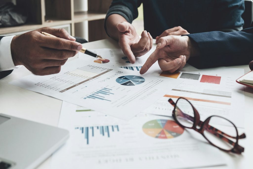 Zwei Personen diskutieren über Finanz- und Forschungsdaten, die auf mehreren Berichten mit Diagrammen und Statistiken dargestellt sind. Die Unterlagen zeigen Balken- und Kreisdiagramme, die auf einem Schreibtisch liegen, zusammen mit einer Brille und einem Stift. Das Bild symbolisiert strategische Planung und Analyse im Zusammenhang mit der Forschungszulage 2026.