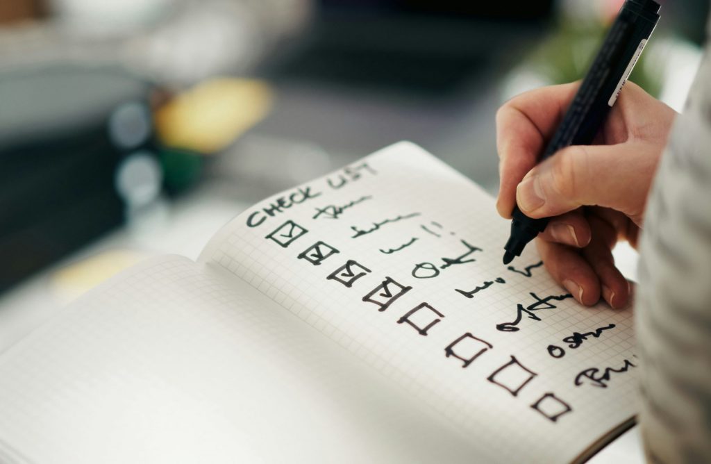 Eine Hand schreibt eine strukturierte Checkliste in ein Notizbuch, um Planung und Bewertungskriterien für gemeinschaftlich entwickelte Geschäftsmodelle systematisch festzuhalten und abzubilden.