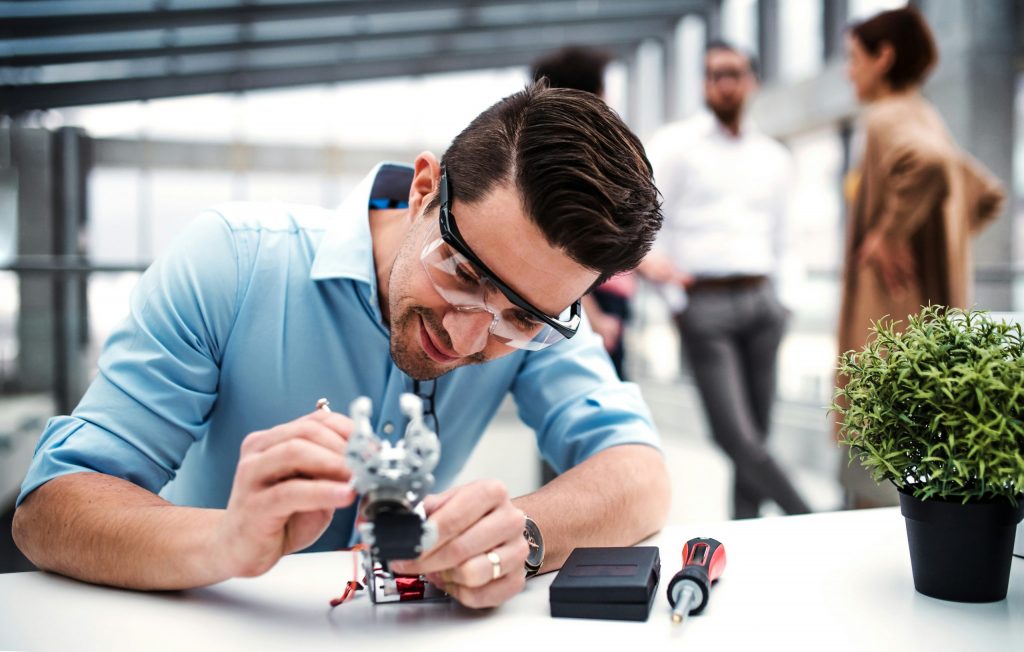 Eine Person arbeitet konzentriert an einem technischen Modell am Schreibtisch, während im Hintergrund weitere Personen im Austausch stehen – ein Sinnbild für gemeinschaftlich entwickelte Geschäftsmodelle und praxisnahe Innovationsentwicklung im Team.