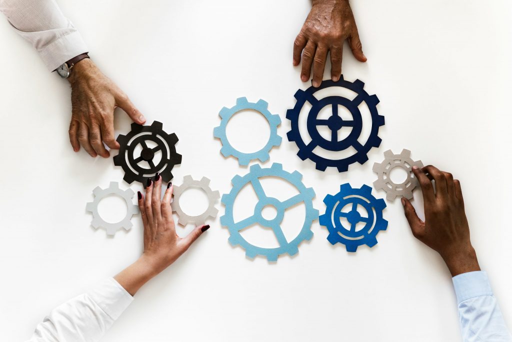 Mehrere Hände positionieren unterschiedlich große Zahnräder in Blau-, Weiß- und Schwarztönen, um gemeinschaftlich entwickelte Geschäftsmodelle visuell darzustellen und ineinandergreifende Zusammenarbeit zu symbolisieren.