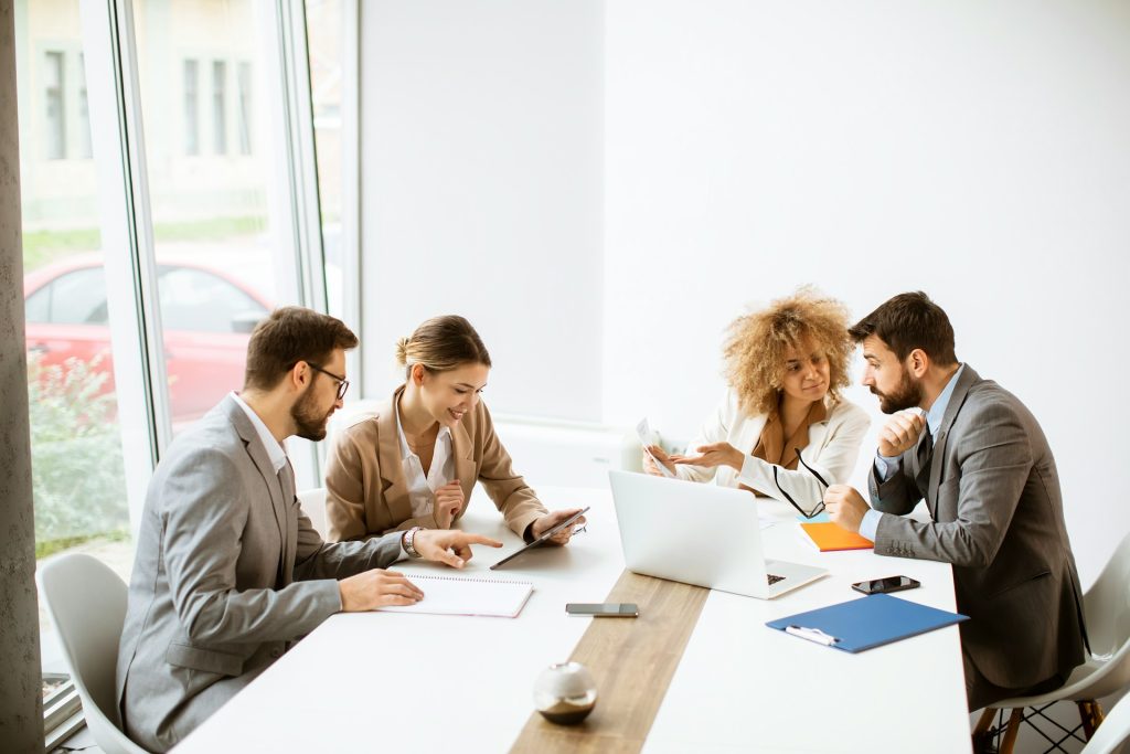 Geschäftsbesprechung mit mehreren Unternehmen an einem Konferenztisch, während zwei Personen per Handschlag eine Zusammenarbeit besiegeln. Das Bild symbolisiert die Zusammenarbeit und Bewertung von Unternehmensstrukturen im Kontext der Forschungszulage für verbundene Unternehmen.