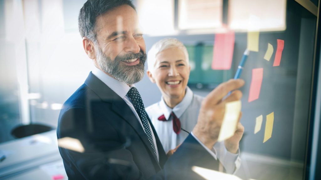Zwei Business Personen schauen mit Freude auf Zettel an der Wand als Symbol für die positive Entwicklung der FuE im Mittelstand durch die Forschungszulage.
