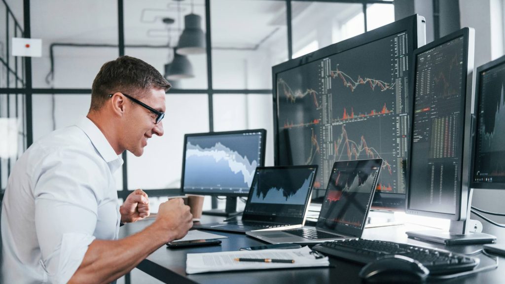 Ein Mitarbeiter sitzt mit Freude vor vielen Monitoren mit Analysen zur Geschäftsentwicklung als Symbol, wie die Forschungszulage in komplexen Unternehmen genutzt wird.