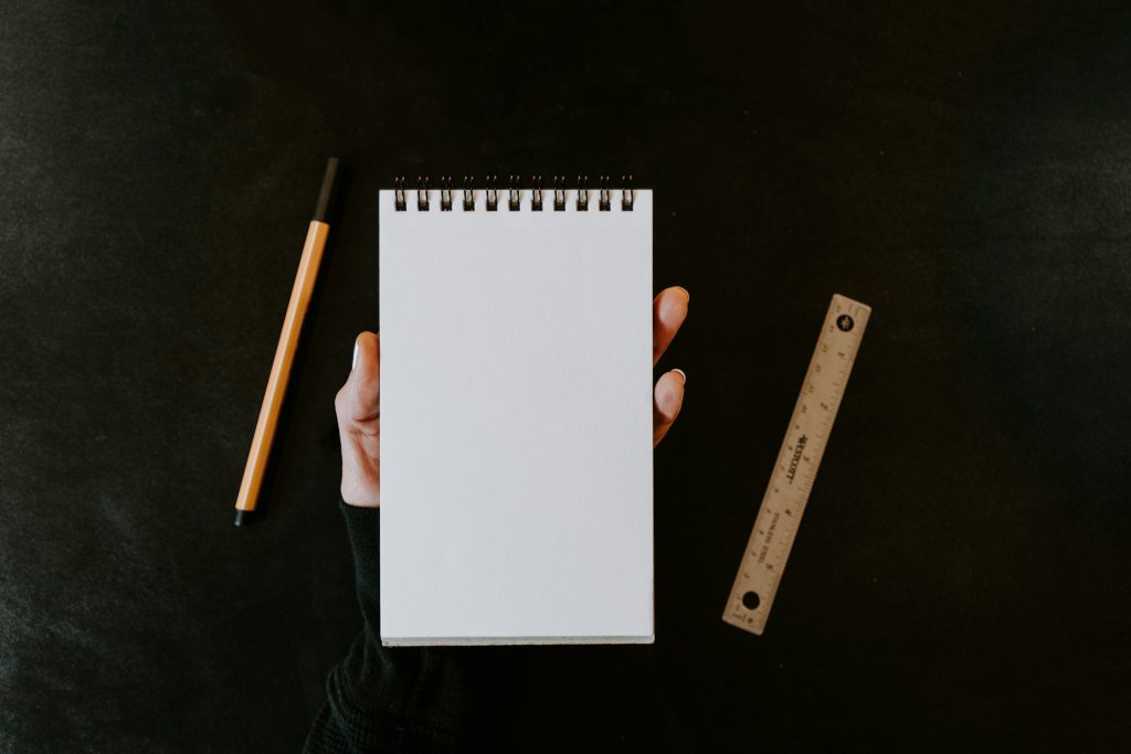 Stundennachweise dokumentieren Arbeitszeiten systematisch: Hand hält einen leeren Notizblock neben Stift und Lineal auf dunklem Schreibtisch.